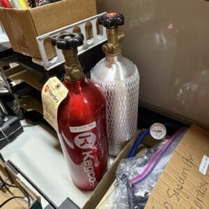 Two gas cylinders (red and silver) with brass regulators and black handwheels sit upright on a cluttered desk next to a pen organizer box with pencils and pens nearby.
