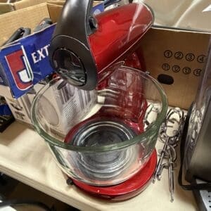 Red stand mixer with a clear glass bowl on a shelf among boxes and cords in a workshop setting.