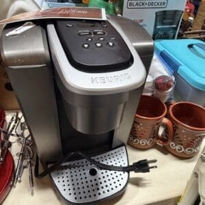 Keurig coffee maker on a kitchen counter with mugs and a box of Skippy peanut butter in the background.