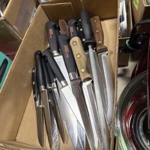 Box of assorted chef knives with black and wooden handles in a cardboard container on a shelf