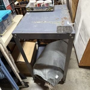 Gray metal utility table on wheels in a cluttered storage room; a gray drawstring bag sits under the lower shelf and a tray of white mugs labeled 'TASSOS' rests on top.