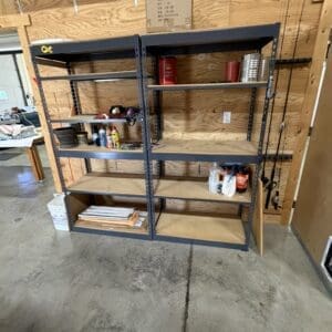 Metal storage shelving unit against a plywood wall in a workshop, with spray cans, tools, and miscellaneous items on the shelves.