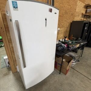 Tall white upright refrigerator in a workshop with wooden walls and a cluttered work table nearby.