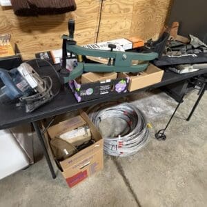 Green metal pipe-bending tool placed on a workbench with cardboard boxes, a coil of metal wire on the floor, and a broom hanging above in a cluttered workshop.