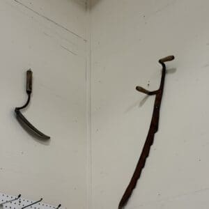 Rusty antique hand tools hanging on a white wall corner: a curved sickle left with a wooden handle and a long, hooked saw on the right.