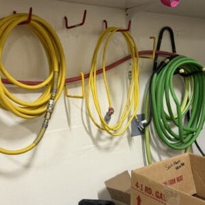 Coiled yellow and green hoses hanging on hooks along a white wall, with a cardboard box and bags on the floor below.