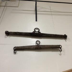 Two weathered wooden lifting bars with iron rings and end hooks mounted on a white wall, likely antique rigging gear in a workshop setting.