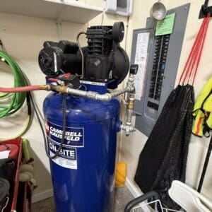 Blue Campbell Hausfeld air compressor with hoses connected in a small workshop corner by a wall-mounted electrical panel.