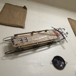 Wooden old-fashioned toboggan sled with metal runners lying on a white floor, with a round black lid nearby.