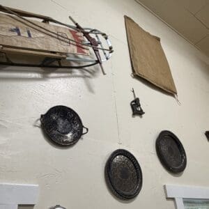 Wall display of three blackened metal pans hanging on hooks, with a worn wooden sled and a cardboard panel visible above on a beige wall.