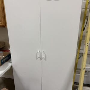White double-door storage cabinet in a workshop, with white curved handles and a base pedestal.