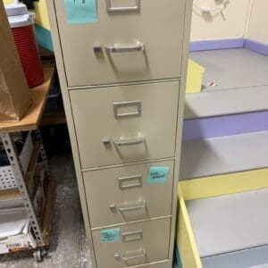 Beige tall metal filing cabinet with several drawers and blue sticky notes attached to two drawers in a cluttered workshop corner.