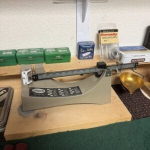 Vintage mechanical OHAUS scale on a wooden reloading bench, surrounded by small green ammo boxes and reloading supplies.