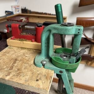 Green manually operated reloading press mounted to a wooden workbench with a teal handle and brass components nearby.