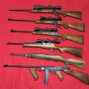 Seven long guns with wooden stocks laid out on a red cloth, several equipped with scopes and bipods (informational image).