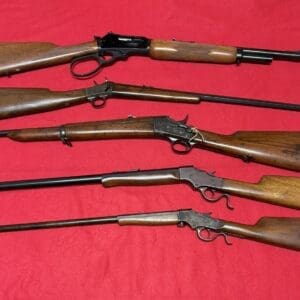 Five vintage rifles with wooden stocks laid on a red surface on top of a fabric background.