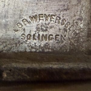 Close-up of a worn metal surface showing a stamped maker's mark, with 'SOLINGEN' partially legible amid patina and scratches.