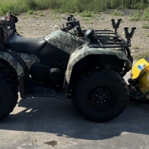 Camouflage all-terrain vehicle (ATV) with large off-road tires, loaded with gear and a yellow front snow plow attachment in front.