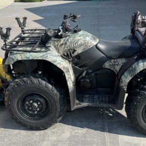 Camouflage ATV with a yellow front snow plow and a gear bag on the rear rack, parked on a driveway.