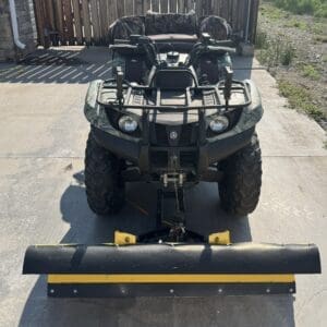 Front view of a camo-pattern ATV with a mounted snow plow on a concrete driveway.