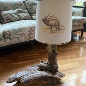 Rustic table lamp with a driftwood base and a shade printed with an elk drawing on a hardwood floor in a living room.