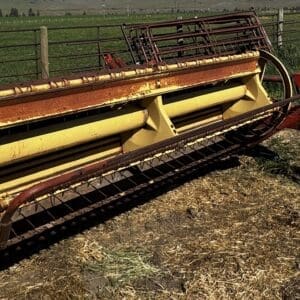 LOT 1 2 - Montana Auctions by Kevin Hill A weathered yellow farm baler sits on dirt near a pasture fence, showing rust and wear in a rural field.