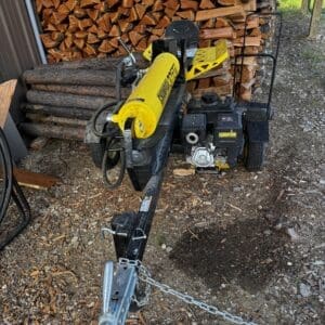 LOT 11 2 - Montana Auctions by Kevin Hill Gas-powered log splitter on a trailer sits in front of a stacked pile of firewood, ready for use or transport, with a tow hitch and safety chain in the foreground.