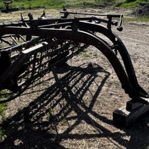 LOT 2 1 - Montana Auctions by Kevin Hill Rusty farming implement with curved metal arms and tines resting on dirt, casting shadows on the ground.