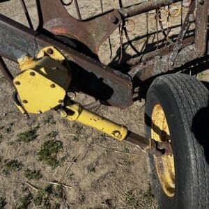 LOT 2 3 - Montana Auctions by Kevin Hill Rusty farm implement frame with a yellow hitch and wheel on dirt ground, showing worn metal and rusted connections.