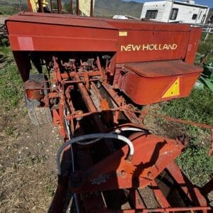 LOT 4 6 - Montana Auctions by Kevin Hill Red New Holland farm implement in a field, showing exposed linkage and hydraulic hoses under a clear blue sky with mountains in the background.