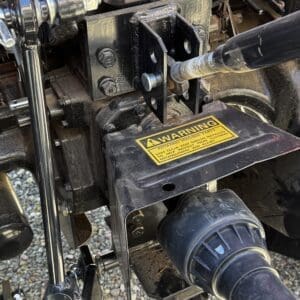 LOT 5 3 - Montana Auctions by Kevin Hill Close-up of a tractor rear hitch and PTO shaft area, showing mounting bolts, a metal bracket, and a yellow warning label about rotating parts.
