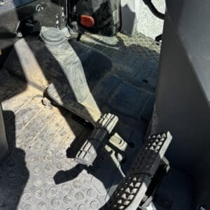 LOT 5 7 - Montana Auctions by Kevin Hill Foot pedals inside a vehicle cab (likely heavy machinery): two large pedals on a textured rubber floor mat, with a dusty surface nearby and an orange lever in the background.