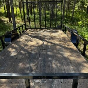 LOT 6 2 - Montana Auctions by Kevin Hill Wooden flatbed trailer in a forest, with a metal gate at the back and surrounding trees and greenery.