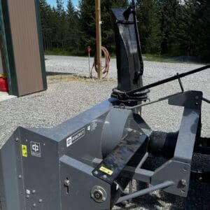 LOT 7 1 - Montana Auctions by Kevin Hill Heavy gray IronCraft skid-steer attachment on gravel, featuring a hydraulic arm and rotating chute, parked beside a shed with trees in the background.