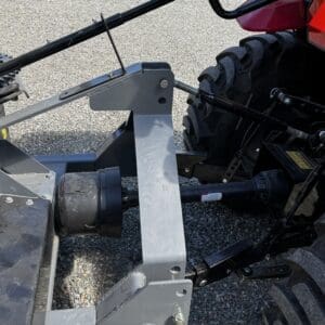 LOT 7 3 - Montana Auctions by Kevin Hill Rear three-point hitch on a tractor with hydraulic lift and PTO shaft, viewed from the side on a gravel surface, with a large tire in the background.