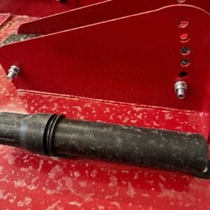 LOT 9 5 - Montana Auctions by Kevin Hill Close-up of red metal machinery with multiple drilled holes, bolts, and a black cylindrical rod resting on the surface.
