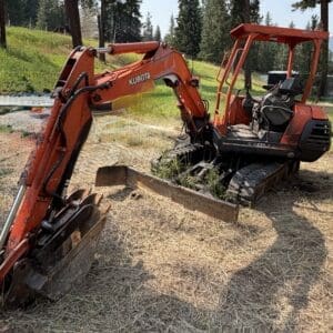 1 1 - Montana Auctions by Kevin Hill Orange Kubota compact excavator with bucket resting on the ground in a dry grassy field, trees in the background.