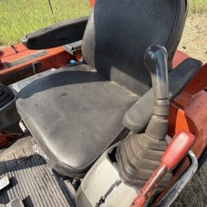1 1 - Montana Auctions by Kevin Hill Operator seat and joystick controls of a red construction tractor, showing a black cushioned seat, right-side joystick, and a red lever in a dusty cab area.