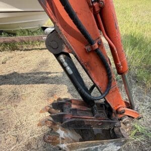 1 2 - Montana Auctions by Kevin Hill Close-up of an orange excavator arm and rusty bucket resting on dry grass, hydraulic hoses visible along the boom.