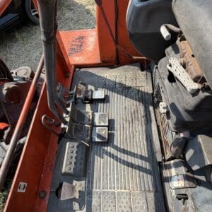 1 2 - Montana Auctions by Kevin Hill Operator's footwell of an orange tractor with multiple pedals and a worn rubber floor mat.