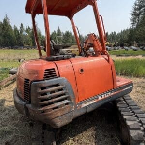 1 6 - Montana Auctions by Kevin Hill Orange Kubota mini excavator with tracks, parked in a dirt lot; boom raised, outdoors.