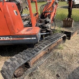 1 7 - Montana Auctions by Kevin Hill Orange mini excavator on a grassy field, showing tracked undercarriage and a rusty bucket attachment ready for work.