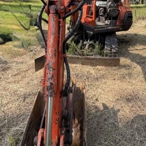 1 8 - Montana Auctions by Kevin Hill Close-up of an orange mini excavator bucket and hydraulic arm resting on a dry grassy field with trees in the background.