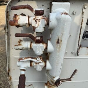 10 1 - Montana Auctions by Kevin Hill Close-up of a rusting gas valve manifold with four valve levers on an outdoor metal unit, showing brown corrosion and peeling paint.
