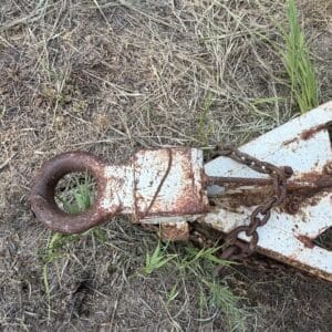 10 3 - Montana Auctions by Kevin Hill Rusty trailer hitch with a large circular eyelet and chain on dry grass and dirt.