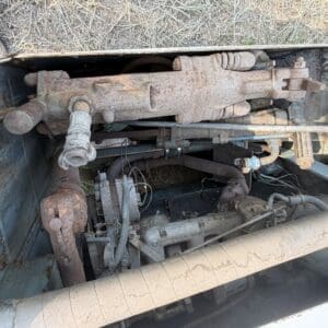 10 3 - Montana Auctions by Kevin Hill Rusty, dusty engine bay components with exposed linkages, hoses, and a large rusted lift arm resting across the area under a vehicle.