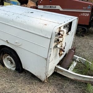 10 4 - Montana Auctions by Kevin Hill White utility trailer/generator with exposed valves at the end, rusted and placed on a trailer hitch in a dry grassy area; a red Ditch Witch machine is in the background.