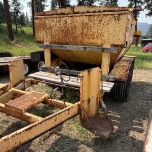 11 2 - Montana Auctions by Kevin Hill Rusty yellow grain trailer with a tilted hopper on a wheeled trailer in a grassy yard with trees in the background.