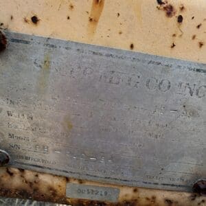 11 6 - Montana Auctions by Kevin Hill Close-up of a weathered metal plaque attached to a rusty beam outdoors; the engraved text is mostly faded with rust spots and a small serial plate at the bottom.