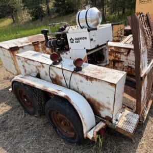 12 1 - Montana Auctions by Kevin Hill Rusted white industrial trailer with a two-tire axle, carrying a compact machine labeled “CUSHION CUT” and exposed hoses, outdoors on grass.